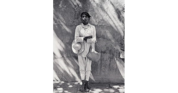 A black-and-white photograph of a dark-skinned man in light-colored shirt and pants, holding a hat under one crossed arm and a bag on the other, leaning against a wall.