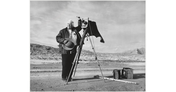 A black-and-white photograph of an older White woman holding a cowboy hat and standing next to a large format camera on a dirt road.