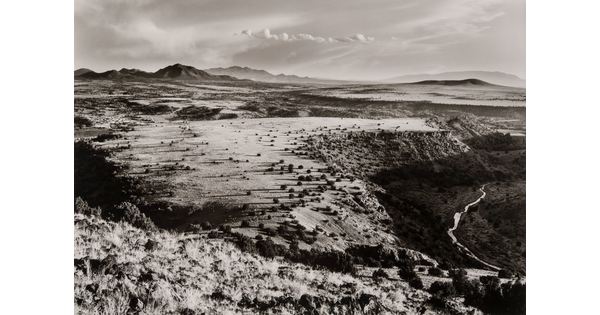 A black-and-white photograph of the top of a mesa with a winding river below and mountains in the distance.