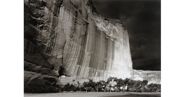 A black-and-white photograph of a sheer cliff face at night with a small ruined structure near the bottom, and trees on the plain in front of the cliff.