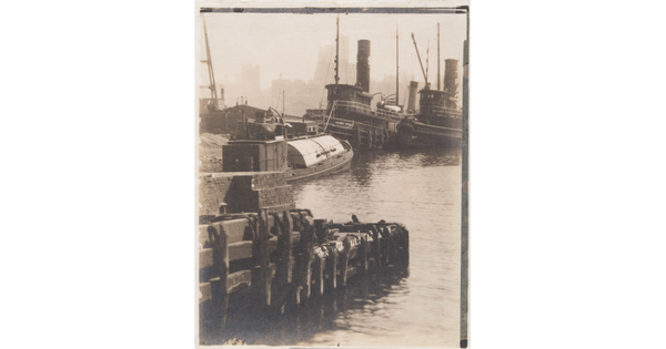 A black-and-white photograph of a narrow wharf with boats alongside and tall buildings in the distance.