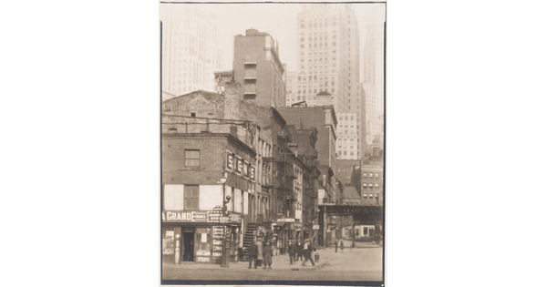 A black-and-white photograph of people on a street corner near three- and four-story buildings with skyscrapers in the background.