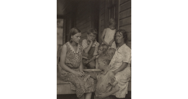 A black-and-white photograph of two White women and several children on the porch of a clapboard house.