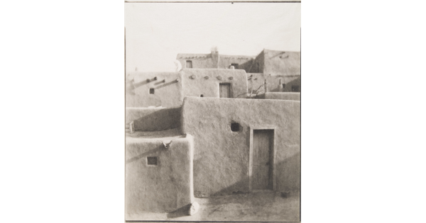 A black-and-white photograph of a multi-level adobe pueblo with wood doors in some of the walls.