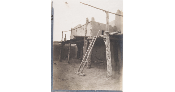 A black-and-white photograph of a multi-level adobe pueblo with a wood ladder and wood framing supporting covered porches.