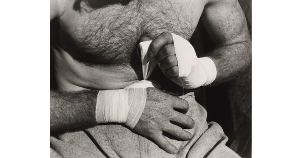 A black-and-white, close-up photograph of a light-skinned, shirtless boxer wrapping his hands with tape.