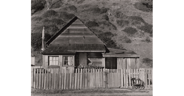 A black-and-white photograph of a small clapboard house behind a picket fence with a chair next to the gate.