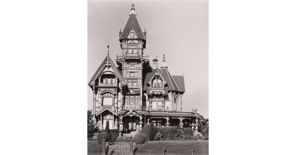 A black-and-white photograph of a large, multi-story, elaborate Victorian house with turrets and a wrap-around porch on a sunny day.