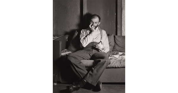 A black-and-white portrait photograph of a bearded White man seated on a sofa with one hand up to his mouth, the other holding a teacup over his lap.