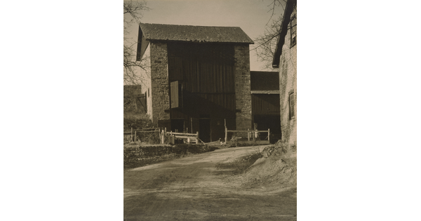 A black-and-white photograph of a dirt road leading to a multi-story stone-sided structure with a wood facade.