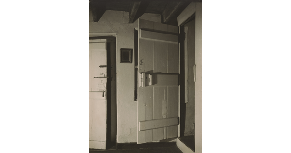 A black-and-white photograph of an interior with light-colored plaster walls and two wood doors: one mostly closed to the left, and the one on the right open.