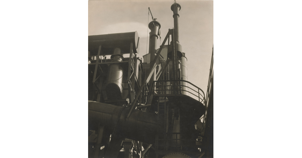 A black-and-white photograph of the exterior of a factory taken from the ground looking up toward the top of its stacks.