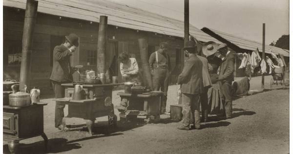 A black-and-white photograph of a group of people, mostly men in bowler hats, standing around outdoor stoves lined up in front of a row of low, long buildings.