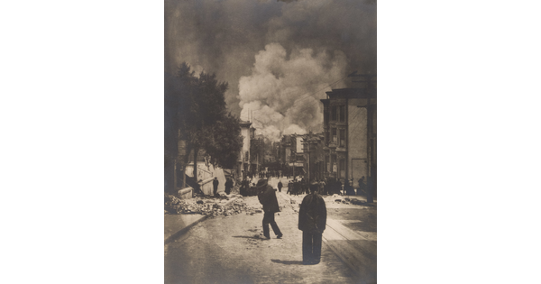 A black-and-white photograph of people standing in a street near piles of rubble and billowing smoke in the distance.