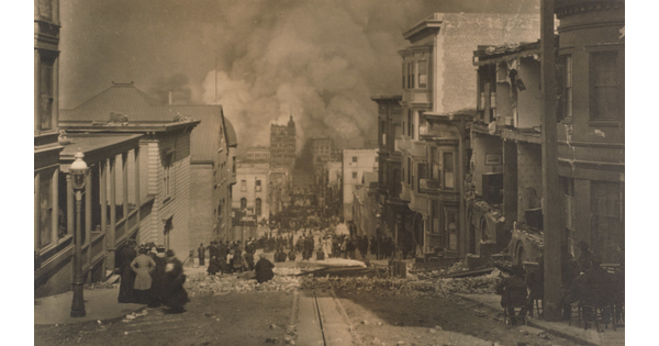 A black and white photograph of people standing in a street filled with rubble from ruined buildings on either side as smoke billows over buildings at the end of the street.
