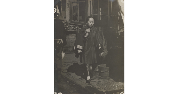 A black-and-white photograph of an woman with Asian features wearing a billowing tunic over pants walking down a commercial street.