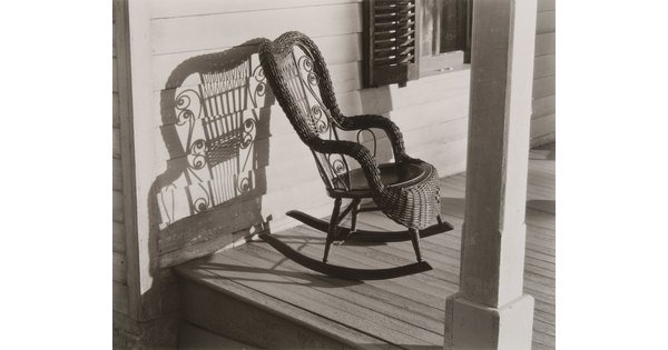 A black-and-white photograph of a decorative wicker rocking chair on a porch that casts a stark shadow on the clapboard behind it.