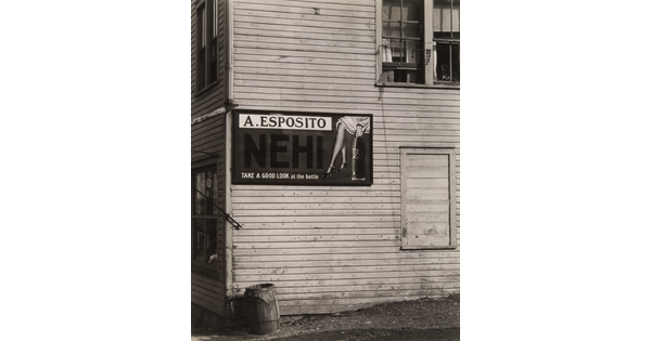 A black-and-white-photograph of the side of an old, clapboard building with a large sign advertising a soft drink.