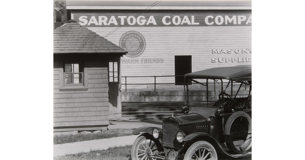 A black-and-white-photograph of a 1920's-style automobile parked in front what looks like a warehouse that is next to railroad tracks.