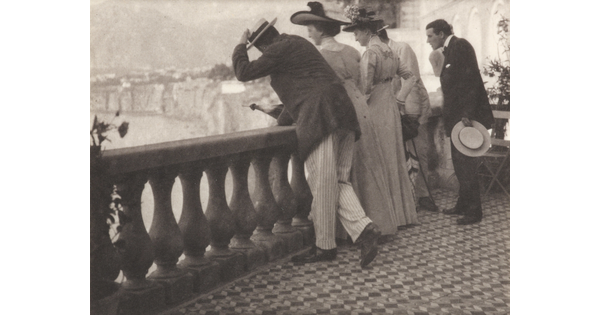 A black-and-white photograph of a group of well-dressed people looking over a balcony with a rugged coastline in the distance.