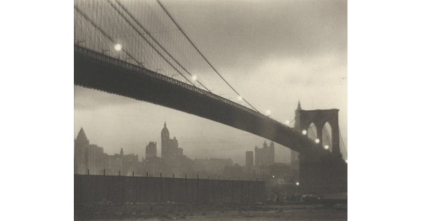 A sepia-toned photograph of a long bridge with a foggy cityscape in the background at nighttime.