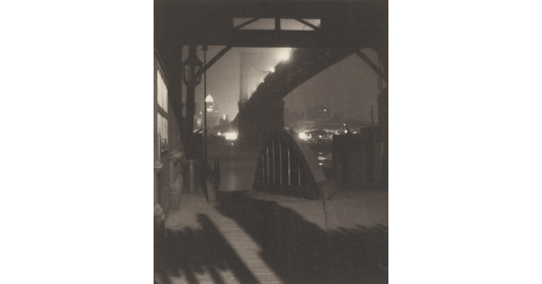 A black-and-white photograph of a dock underneath a large suspension bridge illuminated by street lights.