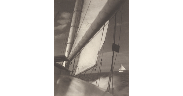A black-and-white photograph looking down the boom of sailboat and sail at another sailboat on the sea in the distance.