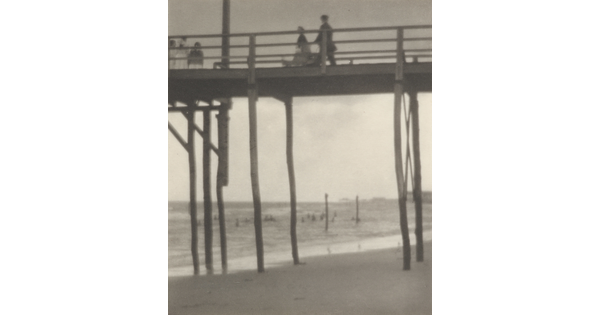 A black-and-white photograph looking up at people walking on a tall pier that sits over a beach.