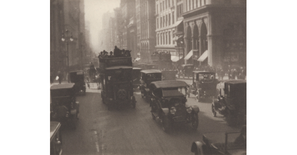 A black-and-white photograph of early 20th-century cars, busses, and wagons pulled by horses on a busy city street.