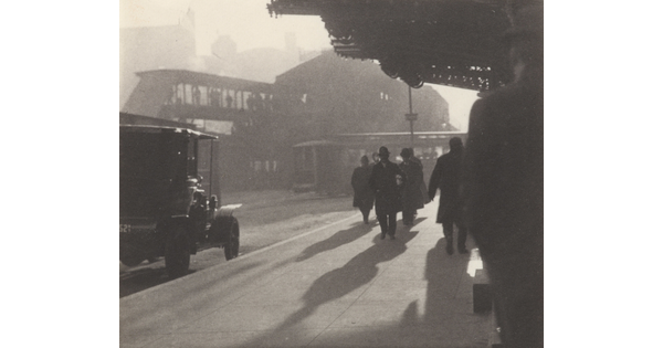 A black-and-white photograph of people walking along the sidewalk of a city street as the sunlight casts long shadows.