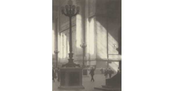A black-and-white photograph of people walking through a sunlit plaza with towering light posts and columns.