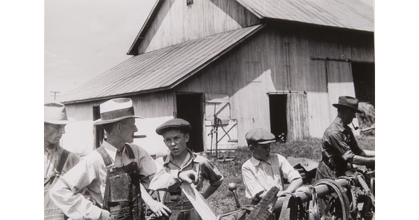 A black-and-white photograph of White men and boys dressed in farming clothes arranging items on a hitching post outside of a barn.