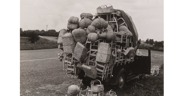 A black-and-white photograph of the back of a truck overflowing with wooden chairs and baskets.