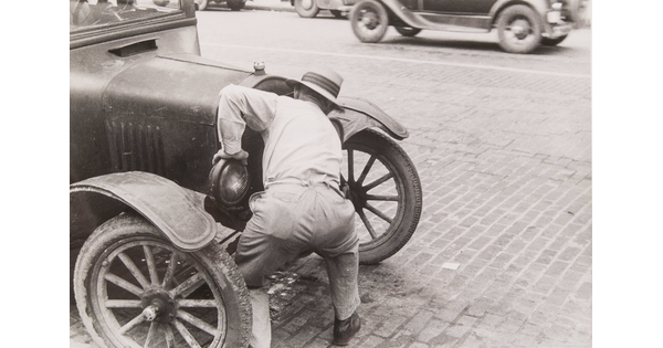A black-and-white photograph of a man bent over the front of an early 20th-century car.