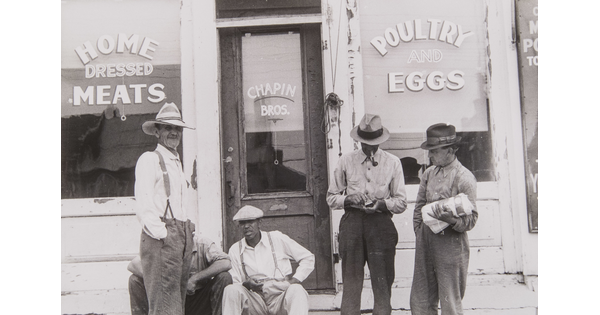 A black-and-white photograph of a group of men, all dressed in work clothes and wearing hats, sitting and standing in front of a store advertising meat, poultry, and eggs.