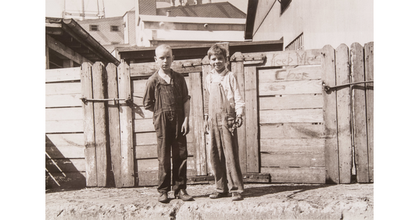 A black-and-white photograph of two White boys dressed in baggy overalls standing in front of a wood fence.