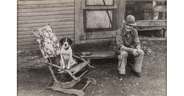 A black-and-white photograph of an elderly man in ragged clothing sitting on a wood stoop as a dog sits in a wood rocker next to him.