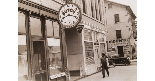 A black-and-white photograph of a Black man walking down a street lined with store fronts, one of which has a large clock face in the foreground.