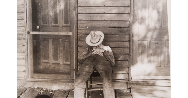 A black-and-white photograph of a man sleeping in a chair, a hat pulled over his face, on the porch of a wood-framed home.