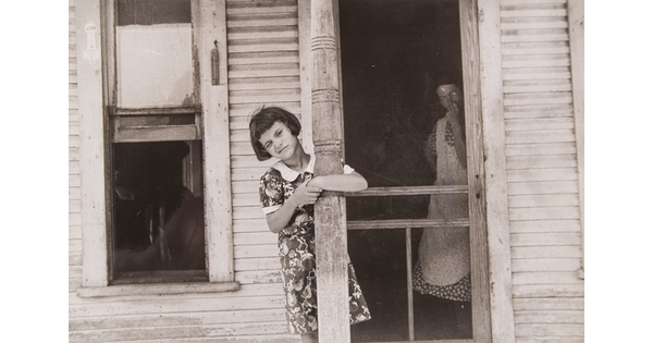 A black-and-white photograph of a young White girl leaning on a porch post as a White woman in a dress and apron stands just inside a screen door.