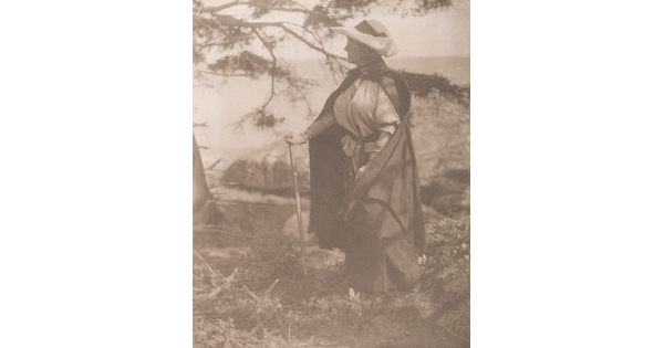 A sepia-toned photograph of a White woman walking in the woods with a walking stick, a large hat, and a cloak.