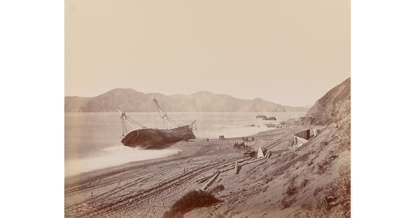 A black-and-white photograph of a large boat tipped on its side in the water near a sandy shore.