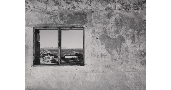 A black-and-white photograph of a concrete wall with a window that reflects an open landscape with mountains in the distance.