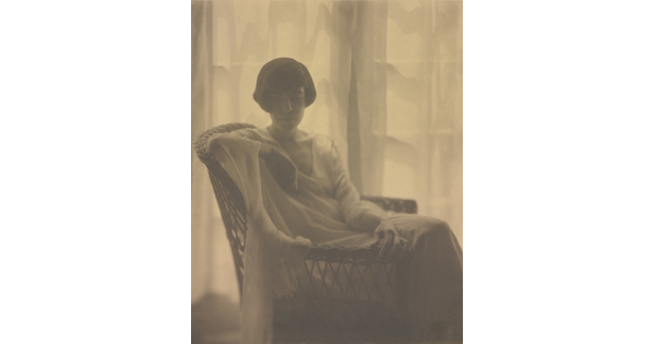 A sepia-toned portrait photograph of a White woman with a bob haircut, wearing a flowing dress and seated in a wicker chair, backlit by sun coming through gauzy curtains.