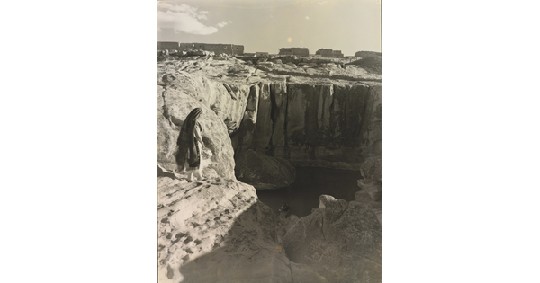 A black-and-white photograph of a woman with a long headscarf on a cliff, overlooking a water hole at the bottom.