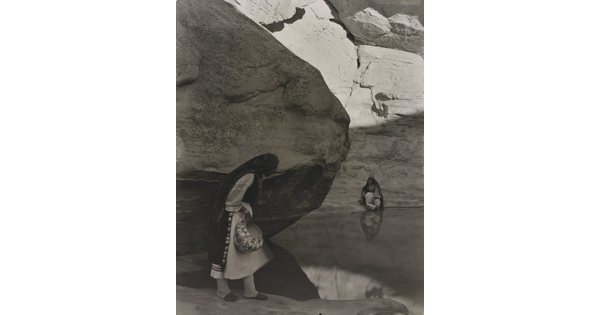 A black-and-white photograph of a woman with a long headscarf standing at the edge of a water hole surrounded by boulders and rock walls.
