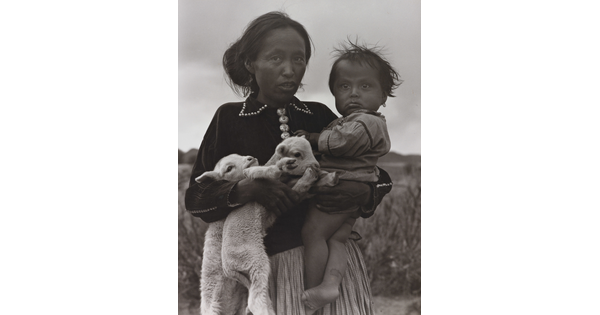 A black-and-white portrait photograph of a Native American woman holding a toddler on one hip and two lambs in her other arm.