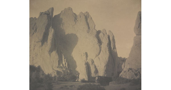 A sepia-toned photograph of tall and broad rock formations with scrubby vegetation at the base of the rocks.