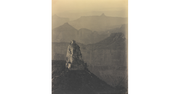 A sepia-toned photograph of rock walls and cliffs fading into the distance; a single rock tower and scrubby vegetation in the foreground.