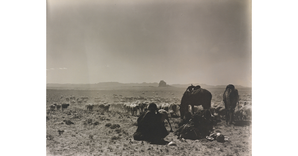 A black-and-white photograph of a man seated on the ground as two saddled horses graze with a herd of sheep on a plain with mountains in the distance.